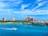 A scenic view of a vibrant waterfront featuring a large resort with multiple pink-colored buildings surrounded by lush greenery and colorful houses. A small white boat is cruising on the clear blue water, while a cruise ship is visible on the distant horizon under a partly cloudy sky.
