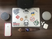 A collection of tech gadgets and accessories displayed on a wooden surface including a laptop covered with various tech-related stickers, two smart speakers, a smartphone, smart watch, and wireless earbuds.