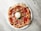 A round pizza with a textured crust, topped with slices of prosciutto, cherry tomatoes, shredded cheese, and a large ball of mozzarella in the center, garnished with green leaves. The pizza is placed on a white plate against a marble surface.