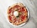 A round pizza with a textured crust, topped with slices of prosciutto, cherry tomatoes, shredded cheese, and a large ball of mozzarella in the center, garnished with green leaves. The pizza is placed on a white plate against a marble surface.