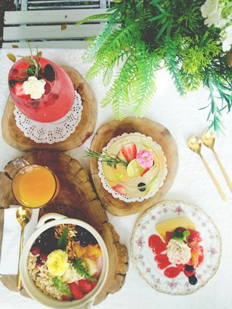 A table is elegantly set with a variety of colorful food and drinks, featuring a pink drink garnished with flowers and berries, a fresh fruit tart, a bowl of granola and fruit, and a plate with dessert covered in berry sauce. Greenery decorates the scene, adding a natural touch to the vibrant display.