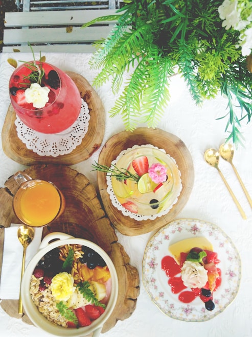 A table is elegantly set with a variety of colorful food and drinks, featuring a pink drink garnished with flowers and berries, a fresh fruit tart, a bowl of granola and fruit, and a plate with dessert covered in berry sauce. Greenery decorates the scene, adding a natural touch to the vibrant display.