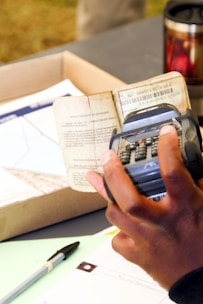 A person is holding a calculator-like device over an open document with printed text. There is a pen and some papers on the table, and a thermal travel mug in the background.