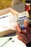 A person is holding a calculator-like device over an open document with printed text. There is a pen and some papers on the table, and a thermal travel mug in the background.