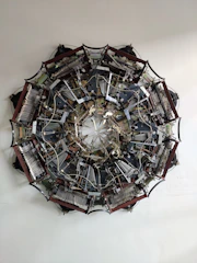 A complex, circular mechanical structure is displayed, composed of numerous interconnected components, gears, wires, and metallic parts arranged symmetrically. The intricate assembly resembles a large, industrial mandala with its numerous parts radiating from the center.