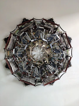 A complex, circular mechanical structure is displayed, composed of numerous interconnected components, gears, wires, and metallic parts arranged symmetrically. The intricate assembly resembles a large, industrial mandala with its numerous parts radiating from the center.