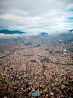 A sprawling cityscape viewed from above, with densely packed buildings and structures. The city is surrounded by mountainous terrain under a slightly overcast sky with clouds partially covering the mountains. A river or waterway snakes through the urban area, adding a natural element to the otherwise tight urban grid.