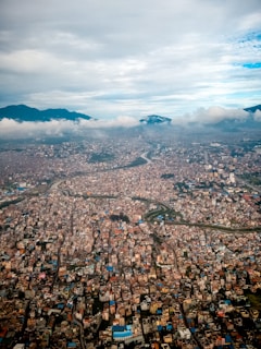 A sprawling cityscape viewed from above, with densely packed buildings and structures. The city is surrounded by mountainous terrain under a slightly overcast sky with clouds partially covering the mountains. A river or waterway snakes through the urban area, adding a natural element to the otherwise tight urban grid.