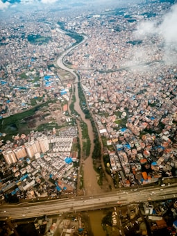 An aerial view of a densely populated urban area with numerous buildings and structures. A river winds through the cityscape, flanked by roads and bridges. Various patches of greenery are visible, interspersed among the urban sprawl. The image is taken from high above, showing extensive detail of the city's layout.