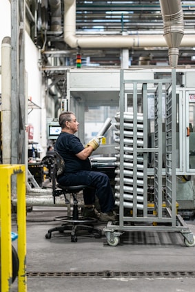 A person wearing work clothes and gloves is seated on a wheeled chair in an industrial setting. They are holding metallic cylindrical objects and are positioned next to a metal rack filled with similar items. The background features machinery, conduits, and an industrial environment.