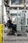 A person wearing work clothes and gloves is seated on a wheeled chair in an industrial setting. They are holding metallic cylindrical objects and are positioned next to a metal rack filled with similar items. The background features machinery, conduits, and an industrial environment.