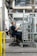 A person wearing work clothes and gloves is seated on a wheeled chair in an industrial setting. They are holding metallic cylindrical objects and are positioned next to a metal rack filled with similar items. The background features machinery, conduits, and an industrial environment.