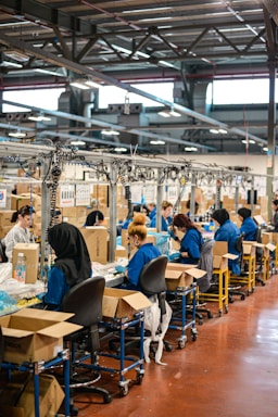 A factory setting with several workers seated at workstations along an assembly line. They are focused on tasks involving sorting or assembling items. The workers are wearing blue uniforms and headgear for safety and hygiene. Cardboard boxes are scattered on and around the tables, and the factory has a ceiling with exposed beams and fluorescent lighting.