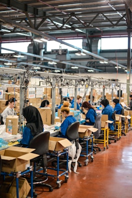 A factory setting with several workers seated at workstations along an assembly line. They are focused on tasks involving sorting or assembling items. The workers are wearing blue uniforms and headgear for safety and hygiene. Cardboard boxes are scattered on and around the tables, and the factory has a ceiling with exposed beams and fluorescent lighting.