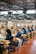 A factory setting with several workers seated at workstations along an assembly line. They are focused on tasks involving sorting or assembling items. The workers are wearing blue uniforms and headgear for safety and hygiene. Cardboard boxes are scattered on and around the tables, and the factory has a ceiling with exposed beams and fluorescent lighting.