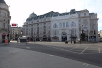 A cityscape featuring a large, historic building with ornate architecture and a dome on the roof. The street in the foreground is mostly empty, with a few pedestrians and a small kiosk visible. A digital billboard is mounted on an adjacent building, and a few traffic lights and lamp posts line the street.