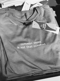 A stack of gray t-shirts with a visible printed message, 'common sense is not that common'. A tag labeled 'THE GOOD PROJECT' is attached to one of the t-shirts. Some price tags are visible in the background.