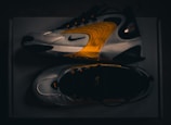 A pair of modern athletic shoes featuring a sleek design with curved lines and a blend of white, black, and vibrant orange hues. The shoes are displayed from a side and top view, highlighting their dynamic style and brand logo.