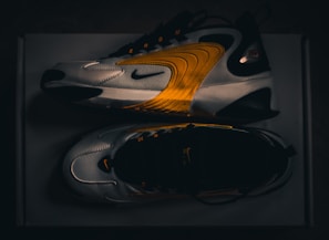 A pair of modern athletic shoes featuring a sleek design with curved lines and a blend of white, black, and vibrant orange hues. The shoes are displayed from a side and top view, highlighting their dynamic style and brand logo.