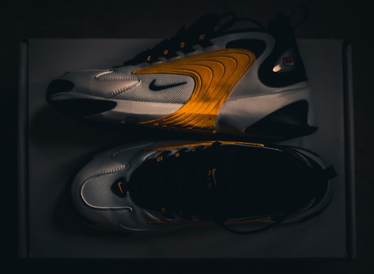 A pair of modern athletic shoes featuring a sleek design with curved lines and a blend of white, black, and vibrant orange hues. The shoes are displayed from a side and top view, highlighting their dynamic style and brand logo.