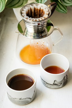 A stainless steel coffee dripper with a white paper filter sits atop a glass carafe containing warm liquid. Two ceramic cups, one with dark and the other with lighter coffee, are positioned in the foreground. The setup is placed on a light-colored surface with green leaves partially visible in the background.