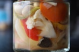 A close-up of a jar filled with a variety of pickled vegetables immersed in liquid. Visible vegetables include a slice of carrot, a piece of cauliflower, and segments of bell pepper. The colors are vibrant and the jar is glass, showcasing the contents clearly.