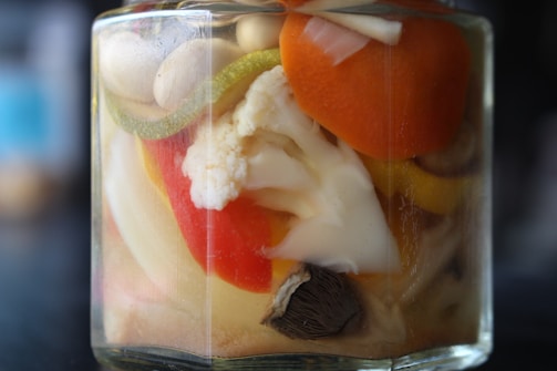 A close-up of a jar filled with a variety of pickled vegetables immersed in liquid. Visible vegetables include a slice of carrot, a piece of cauliflower, and segments of bell pepper. The colors are vibrant and the jar is glass, showcasing the contents clearly.