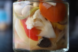 A close-up of a jar filled with a variety of pickled vegetables immersed in liquid. Visible vegetables include a slice of carrot, a piece of cauliflower, and segments of bell pepper. The colors are vibrant and the jar is glass, showcasing the contents clearly.