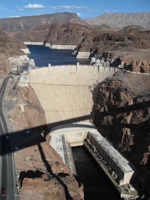 A large concrete dam is set amidst rugged, rocky landscapes with a deep blue reservoir of water behind it. The structure is imposing, with a smooth curved wall and various facilities at its base. There is a road leading up to the dam, flanked by craggy brown mountains under a clear blue sky.