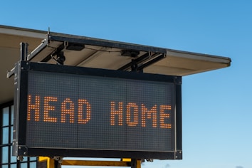 An electronic sign with the words 'HEAD HOME' displayed in bright, orange letters. The sign is mounted on the corner of a building under a clear blue sky.