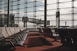 Rows of empty chairs line a spacious airport departure lounge. Large floor-to-ceiling windows offer a view of the tarmac and several jet bridges connecting to airplanes. The scene is tranquil and well-lit by natural daylight, casting shadows across the red carpet.