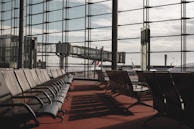 Rows of empty chairs line a spacious airport departure lounge. Large floor-to-ceiling windows offer a view of the tarmac and several jet bridges connecting to airplanes. The scene is tranquil and well-lit by natural daylight, casting shadows across the red carpet.