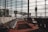 Rows of empty chairs line a spacious airport departure lounge. Large floor-to-ceiling windows offer a view of the tarmac and several jet bridges connecting to airplanes. The scene is tranquil and well-lit by natural daylight, casting shadows across the red carpet.