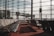Rows of empty chairs line a spacious airport departure lounge. Large floor-to-ceiling windows offer a view of the tarmac and several jet bridges connecting to airplanes. The scene is tranquil and well-lit by natural daylight, casting shadows across the red carpet.