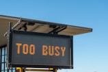 A digital sign displaying the text 'TOO BUSY' in bright orange letters, mounted on a metal frame with a building's roof visible in the background against a clear blue sky.