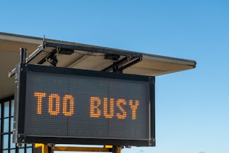 A digital sign displaying the text 'TOO BUSY' in bright orange letters, mounted on a metal frame with a building's roof visible in the background against a clear blue sky.