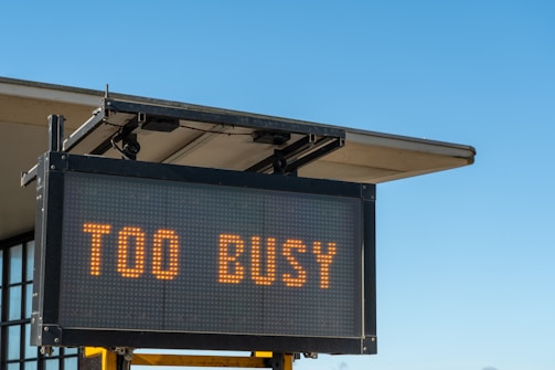 A digital sign displaying the text 'TOO BUSY' in bright orange letters, mounted on a metal frame with a building's roof visible in the background against a clear blue sky.