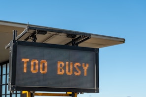 A digital sign displaying the text 'TOO BUSY' in bright orange letters, mounted on a metal frame with a building's roof visible in the background against a clear blue sky.