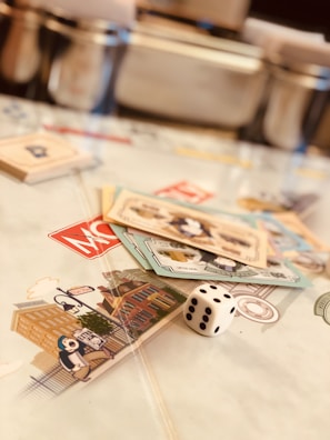 A tabletop setup featuring Monopoly-themed cards and a single die placed on a game board. The cards display various illustrations, while the die shows the number five. The scene is softly lit, creating a warm and inviting atmosphere.