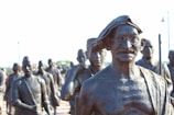 A group of bronze statues with a prominent figure in glasses and traditional attire standing in front. The statues appear to represent historical figures engaged in a march or procession.