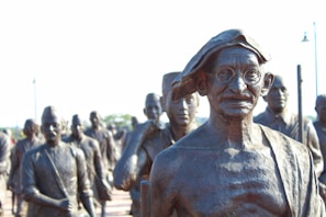 A group of bronze statues with a prominent figure in glasses and traditional attire standing in front. The statues appear to represent historical figures engaged in a march or procession.