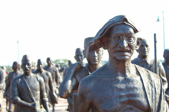 A group of bronze statues with a prominent figure in glasses and traditional attire standing in front. The statues appear to represent historical figures engaged in a march or procession.