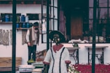 A vintage-looking environment with a mannequin dressed in historical attire and a person in the background. The setting features rustic décor, including fabric patterns and a collection of containers, possibly in a storefront or artistic workspace. The atmosphere appears nostalgic and somewhat cluttered.