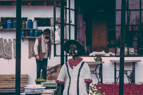 A vintage-looking environment with a mannequin dressed in historical attire and a person in the background. The setting features rustic décor, including fabric patterns and a collection of containers, possibly in a storefront or artistic workspace. The atmosphere appears nostalgic and somewhat cluttered.