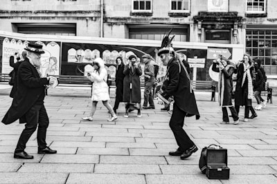 Two performers dressed in traditional attire engage in a lively street performance, attracting the attention of onlookers. One performer plays a musical instrument, while the other is mid-step in a dance. A group of people, some taking photos, stand in the background watching. The setting appears to be an urban street with a building and advertisement panels behind them.