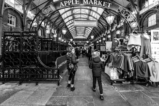 A black and white photograph of the Apple Market, a covered market area with stalls displaying various clothes. The arched metal entrance sign prominently displays the name of the market. People can be seen walking through the market, adding a sense of movement to the scene. The architectural features include a high, vaulted ceiling with hanging lights.