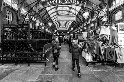 A black and white photograph of the Apple Market, a covered market area with stalls displaying various clothes. The arched metal entrance sign prominently displays the name of the market. People can be seen walking through the market, adding a sense of movement to the scene. The architectural features include a high, vaulted ceiling with hanging lights.
