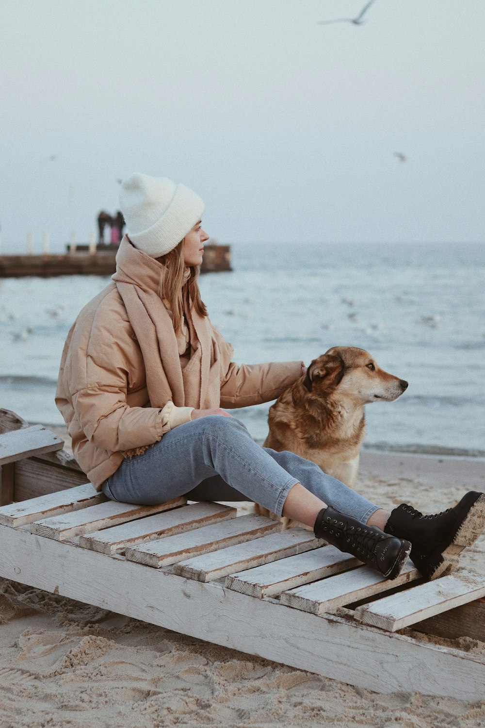 woman in brown jacket and blue denim jeans sitting on brown wooden bench during daytime