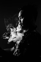 A person is captured in profile against a dark background, with smoke swirling around their face and rising into the air. The lighting is low, creating dramatic contrasts and highlighting the contours of their facial features.