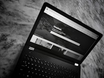 A black laptop is placed on a marble surface. The screen shows the homepage of the Unsplash website, with a header featuring navigation links and a search bar. The section below displays a large background image with text and several smaller images of photography content.